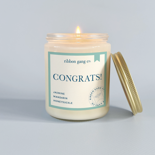 Candle with 'Congrats!' label from Ribbon Gang Co. on a light grey background.