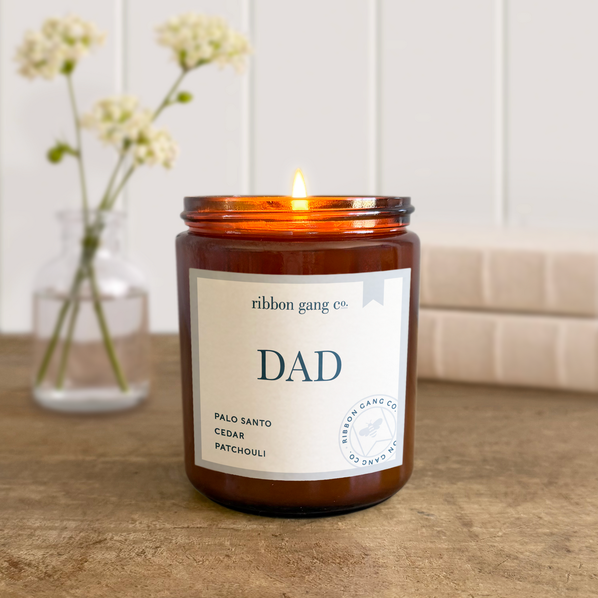 A lit Dad candle in an amber jar on an aged timber tabletop with linen covered books and a jar of flowers stems in the background.