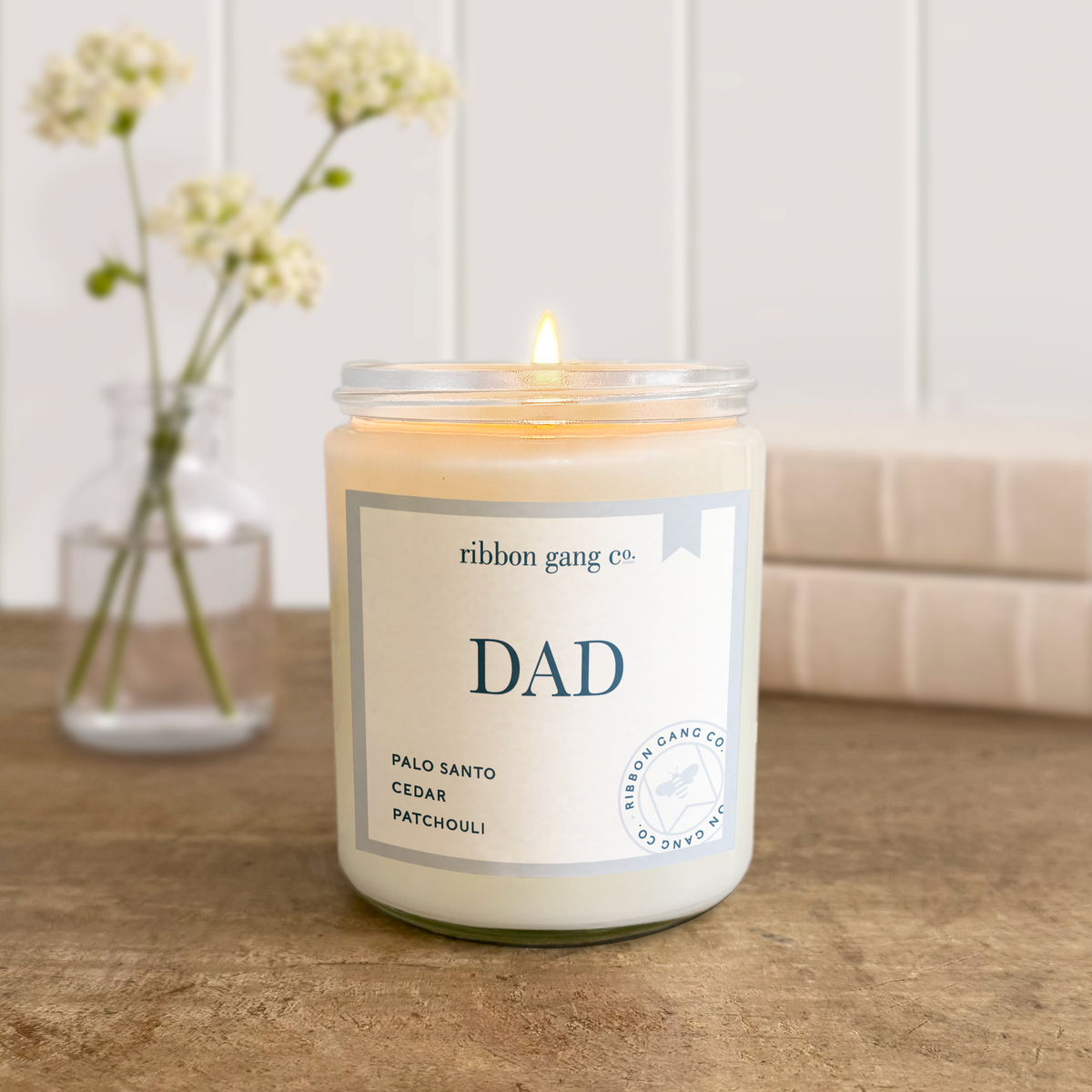 A lit Dad candle in a clear jar on an aged timber tabletop with linen covered books and a jar of flowers stems in the background.