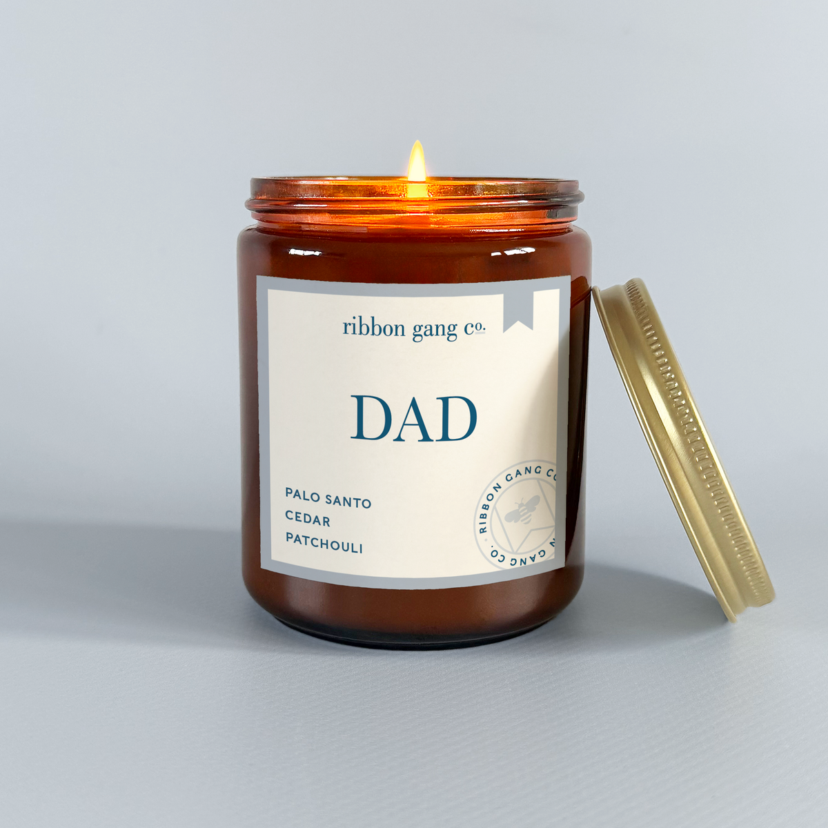 A Dad candle in an amber jar lit with a gold lid resting against it on a pale blue background.