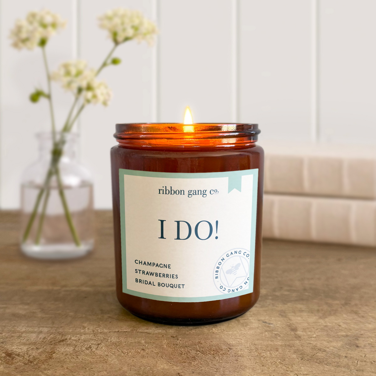 Candle labeled 'I Do!' by Ribbon Gang Co. on a wooden surface with a vase of flowers in the background.